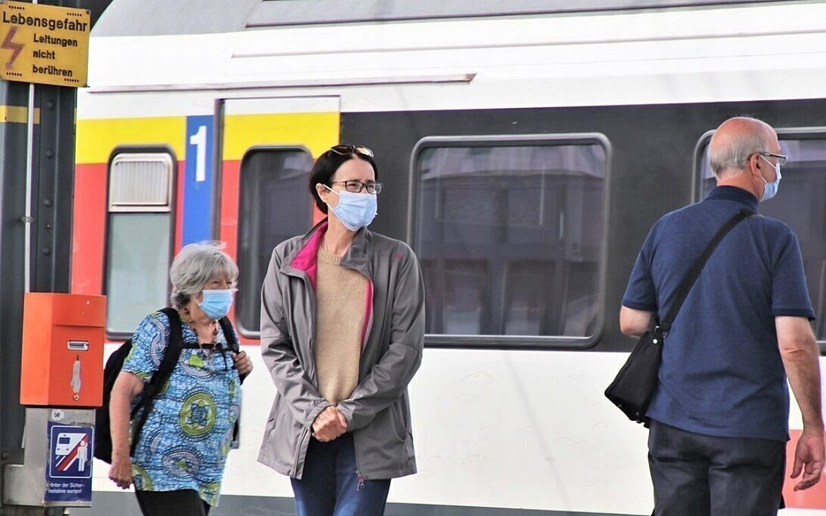 Menschen mit Maske vor einem Zug am Bahnhof Menschen mit Maske vor einem Zug am Bahnhof