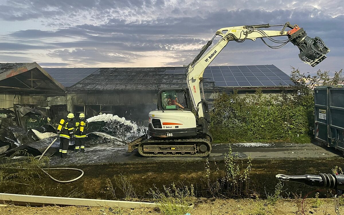 Ein Bagger mit Greifarm steht vor der zerstörten Halle, die Feuerwehr löscht Ein Bagger mit Greifarm steht vor der zerstörten Halle, die Feuerwehr löscht