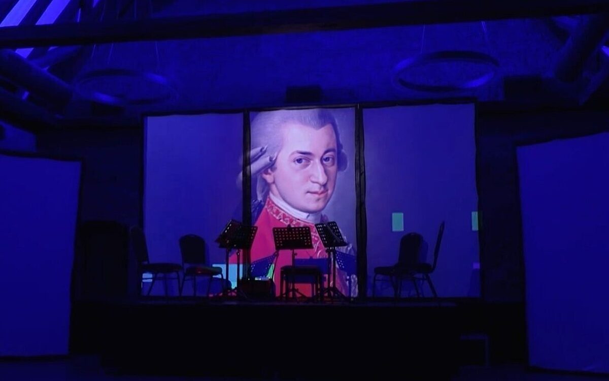 Eine dunkle Bühne bei einem Konzert des Mozarfests. Auf einem Bildschirm erkennt man den Komponisten Mozart Mozartfest