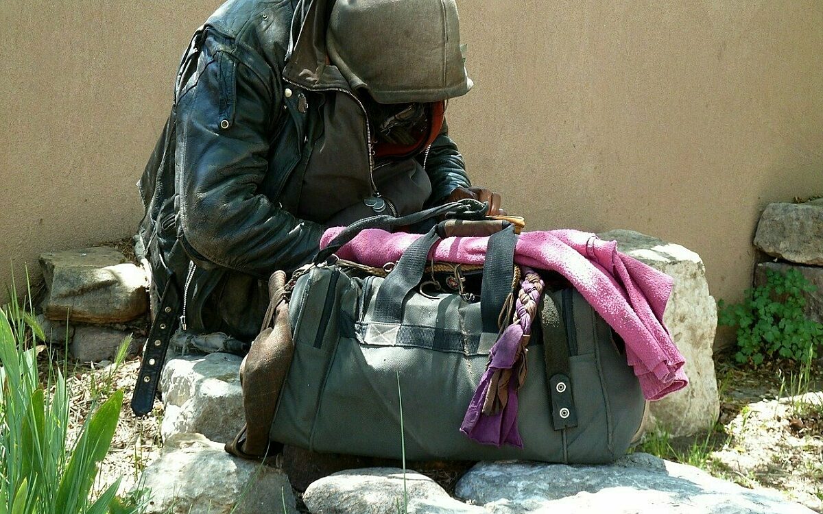 Eine obdachlose Person sitzt mit einer Reisetasche vor einer Mauer Eine obdachlose Person sitzt mit einer Reisetasche vor einer Mauer