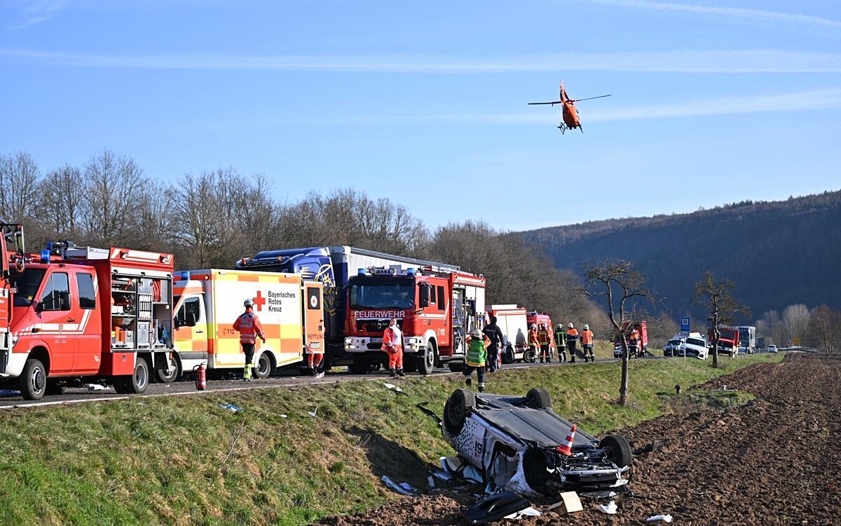 Ein Auto liegt auf dem Dach im Straßengraben, im Hintergrund stehen Rettungsfahrzeuge und ein Hubschrauber startet Ein Auto liegt auf dem Dach im Straßengraben, im Hintergrund stehen Rettungsfahrzeuge und ein Hubschrauber startet